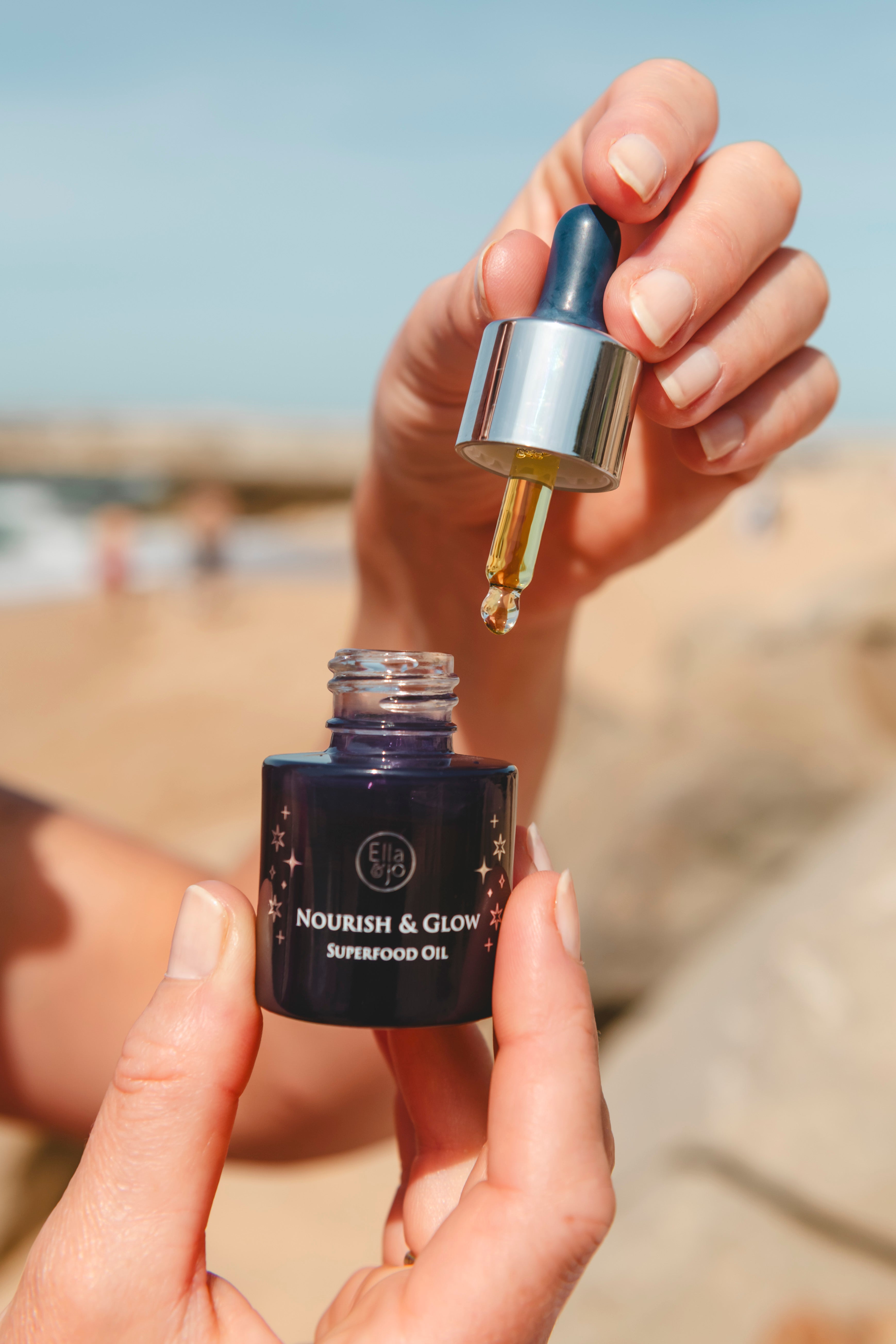 Hand holding a dropper over a bottle labeled 'Nourish & Glow Superfood Oil' with a blurred beach background.